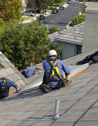 Three workers in safety gear repair or install new roofing materials on a residential rooftop under sunny conditions; nearby rooftops and a street are visible.