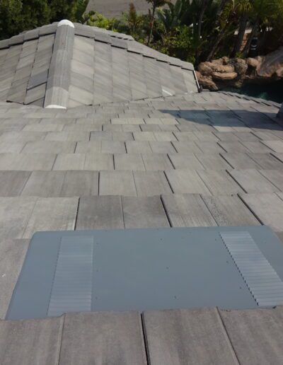 Gray tile roof with a large rectangular metal vent near the foreground; trees and a pool are visible in the background.