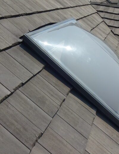 A domed skylight installed on a roof with gray rectangular shingles, showing sunlight reflecting off its surface.