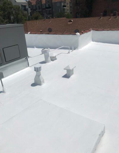 Flat white-painted rooftop with various vents and pipes, bordered by short white parapet walls; residential buildings and trees visible in the background.