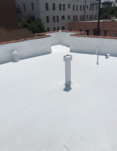 Flat white rooftop with several vent pipes and a parapet wall, surrounded by neighboring buildings and power lines in the background.