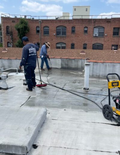 Two workers clean a flat rooftop with hoses and a pressure washer on a sunny day, with buildings visible in the background.